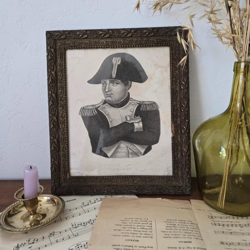 Portrait de Napoléon dans un cadre ancien exposé sur une mise en scène poétique avec partitions anciennes, chandelier et dame-jeanne ambré.