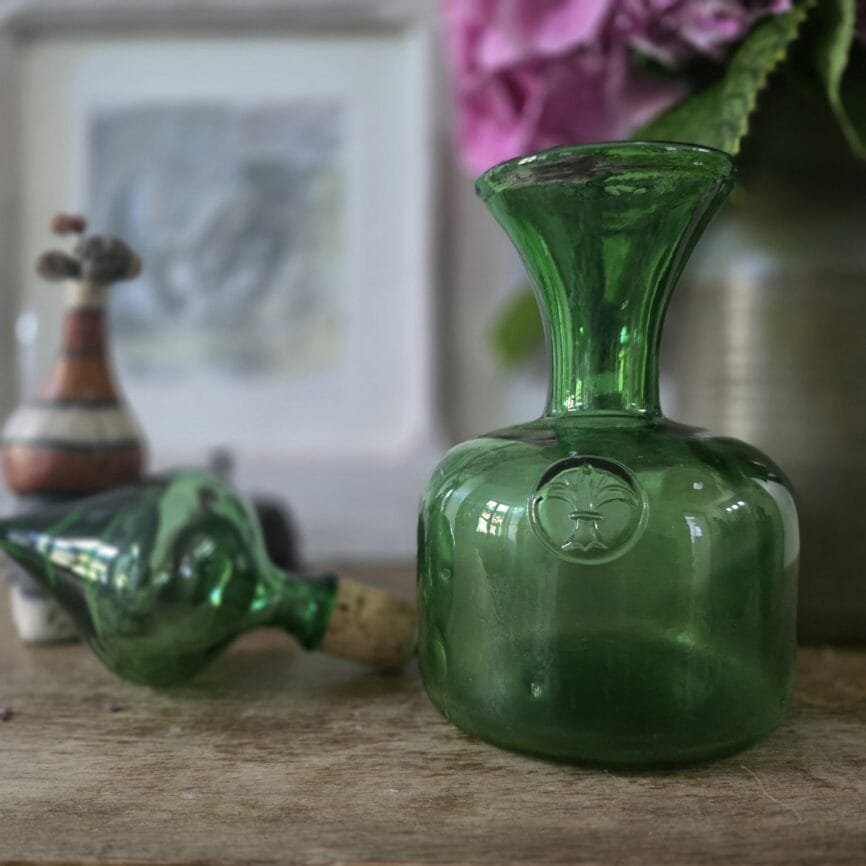 Carafe verte vintage en verre soufflé, bouchon assorti, posée sur un meuble avec fleurs en fond.