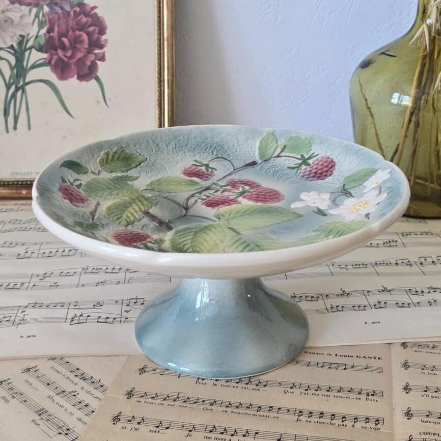 Compotier ancien en barbotine de St Clément, décor framboises et fleurs sur fond bleu, présenté sur partitions anciennes.