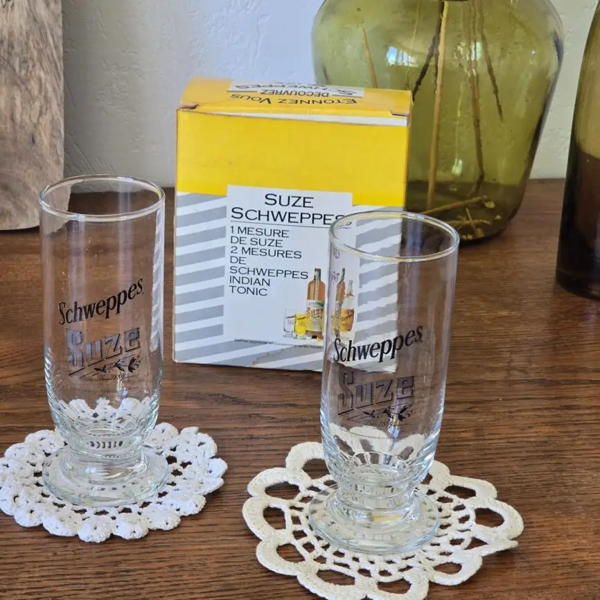 Coffret ouvert avec les deux verres posés sur napperons rétro, bouteille décorative en arrière-plan.