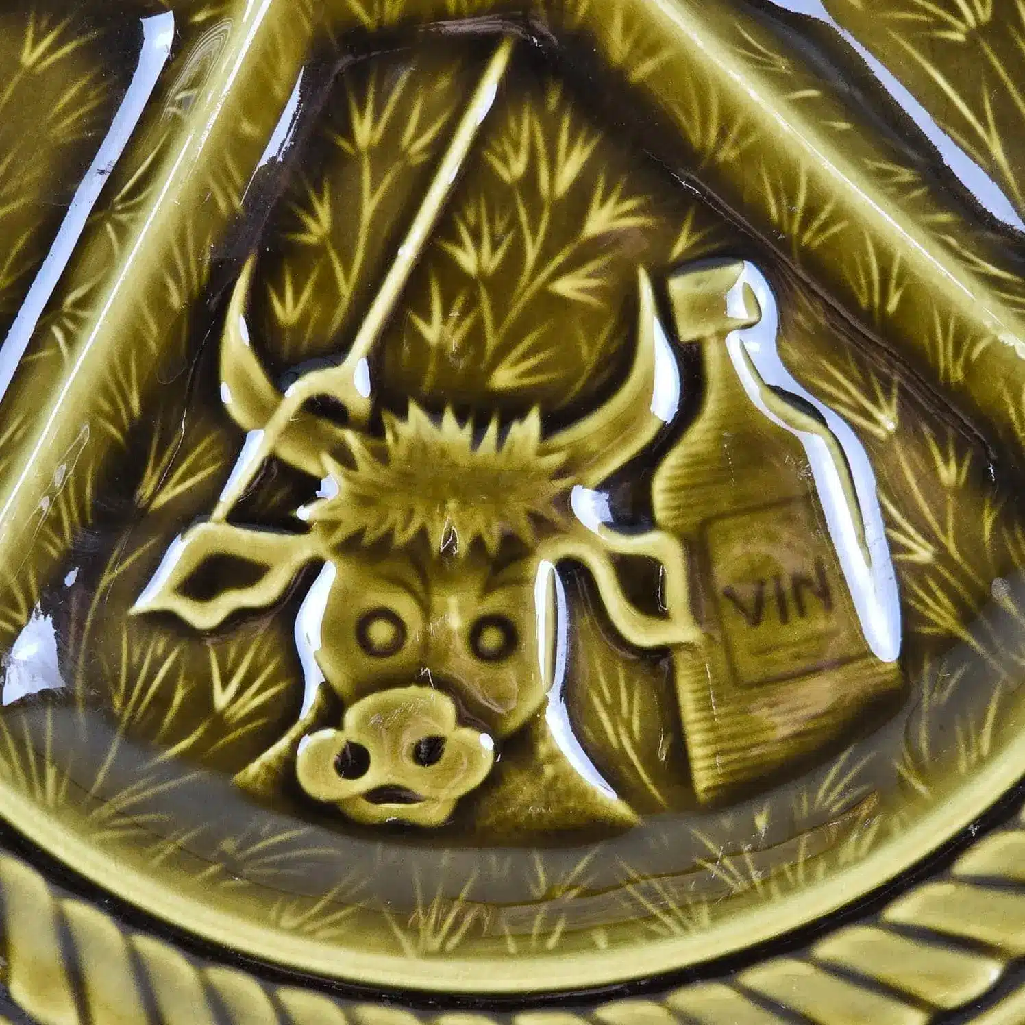 Zoom sur le motif en relief d’une vache et d’une bouteille de vin sur une assiette à fondue Sarreguemines.