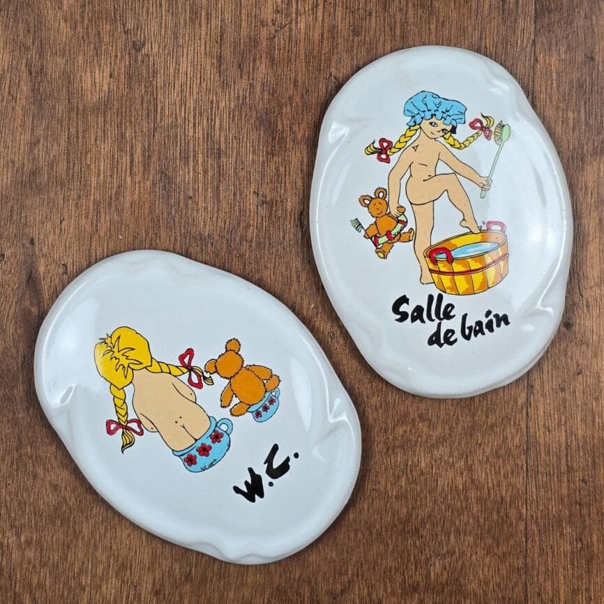 Lot de deux plaques de porte vintage en céramique blanche illustrées d’une fillette aux nattes blondes et de son ours, inscriptions “Salle de bain” et “W.C.” sur fond bois.