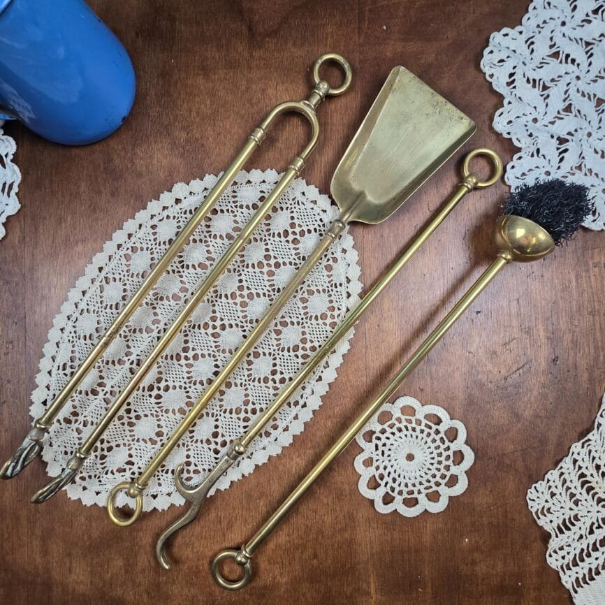 Ensemble d’outils de cheminée anciens en laiton doré posés sur une nappe en dentelle vintage.