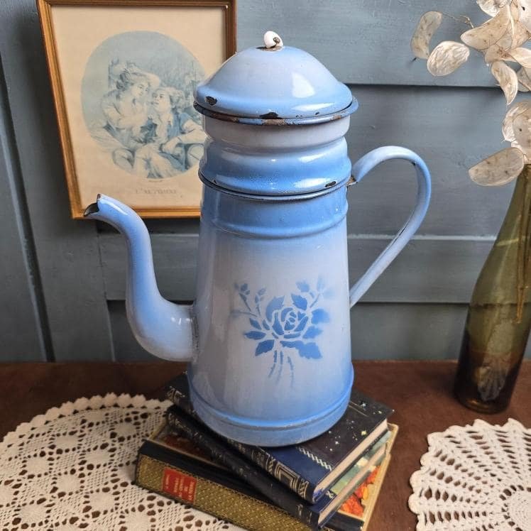 Grande cafetière ancienne émaillée bleue à décor de fleur, posée sur des livres anciens et un napperon crochet, ambiance vintage et douce lumière.