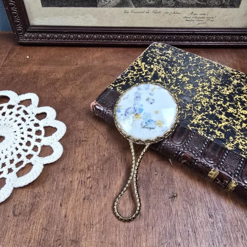 Miroir de sac Limoges posé sur un livre ancien, décor fleuri bleu et jaune.