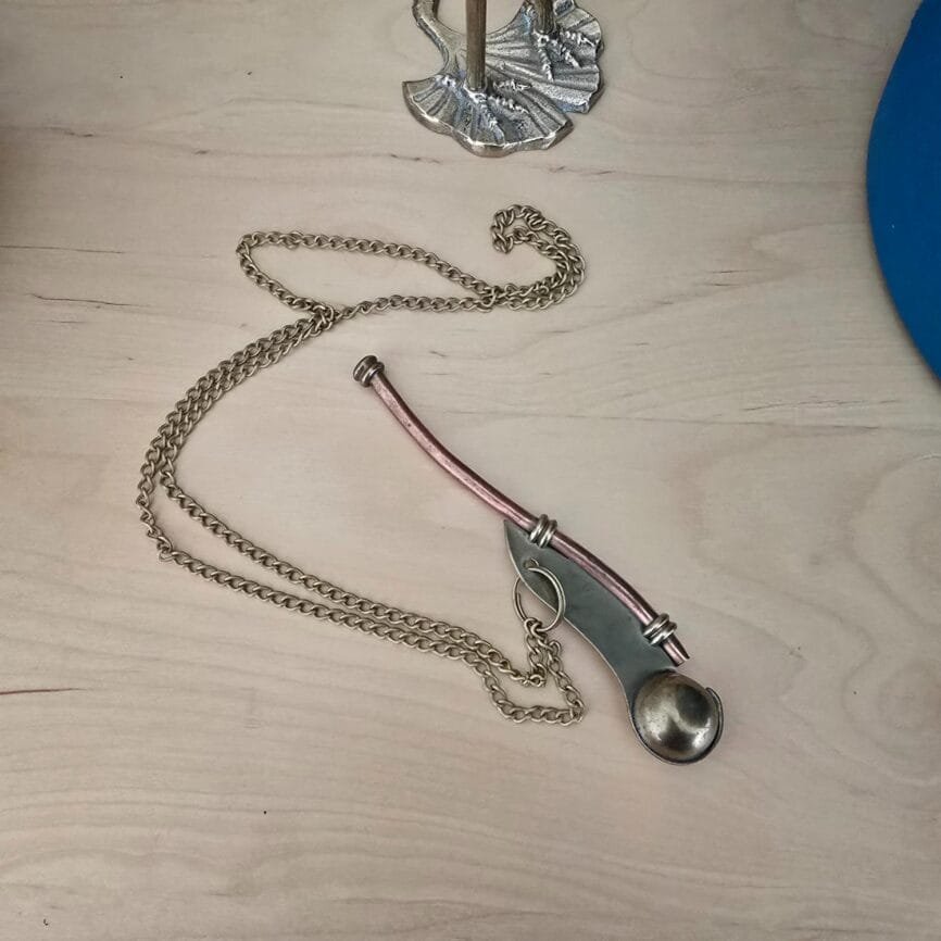Sifflet de bosco ancien en cuivre et laiton posé sur un fond en bois clair, avec sa chaîne métallique. Pièce authentique utilisée autrefois pour les manœuvres en mer.