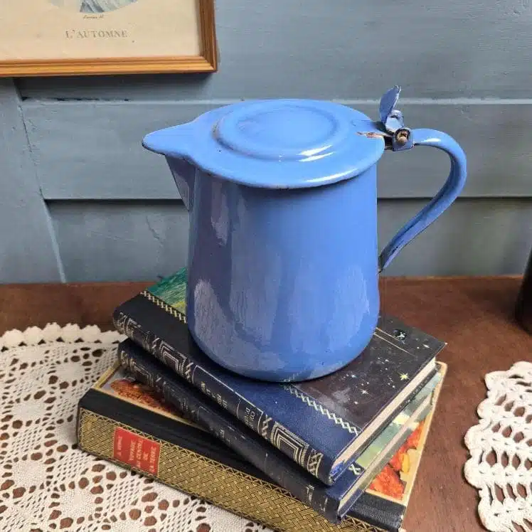 Verseuse ancienne émaillée bleue, petit format rare, posée sur des livres anciens et un napperon crochet, ambiance rétro et douce lumière.