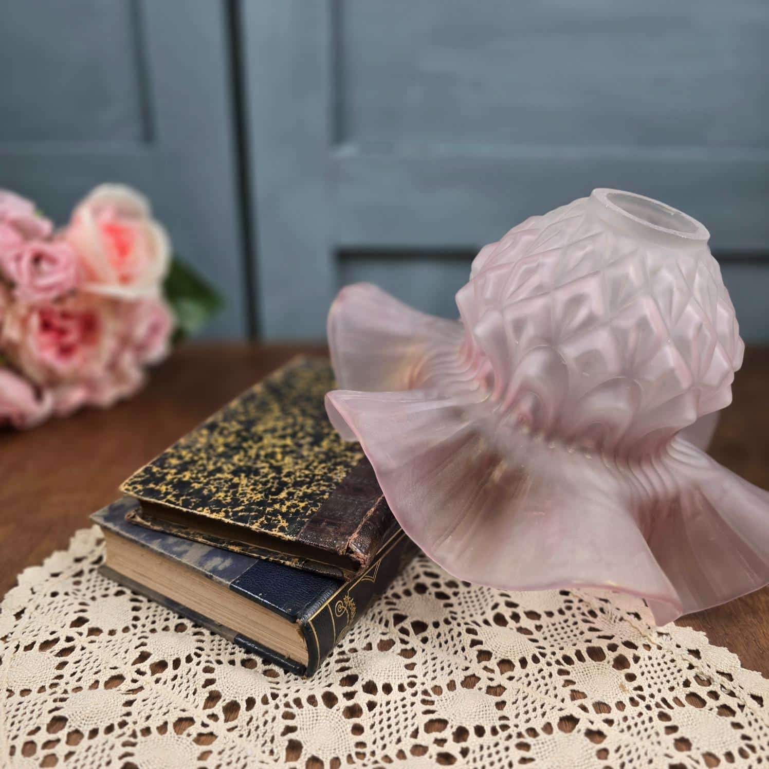 Abat-jour ancien en verre rose satiné, bord ondulé, posé sur des livres sur table en bois et napperon