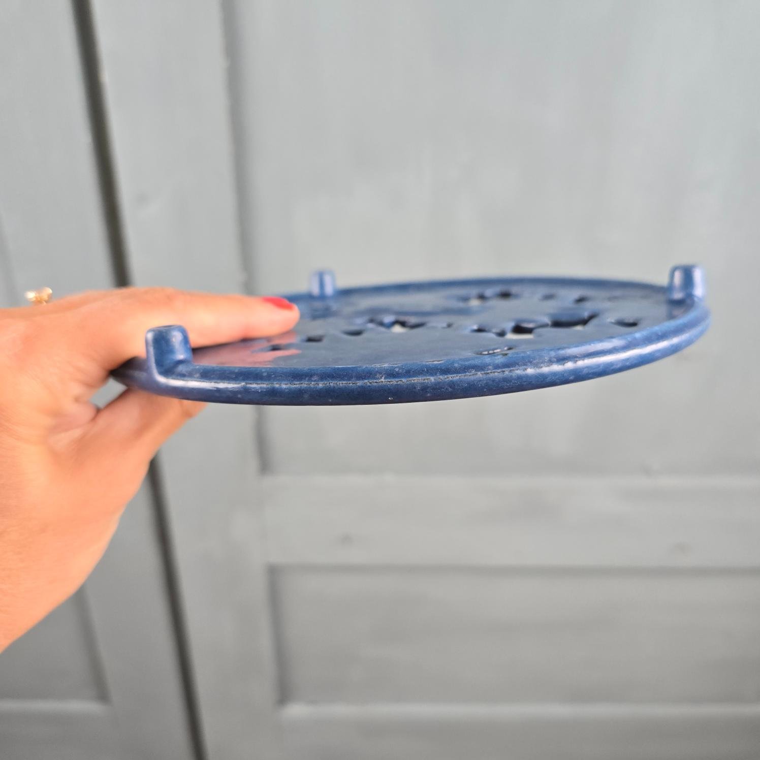 Dessous de plat en fonte émaillée bleue à fleurs Invicta tenu en main de profil pour montrer les petits pieds