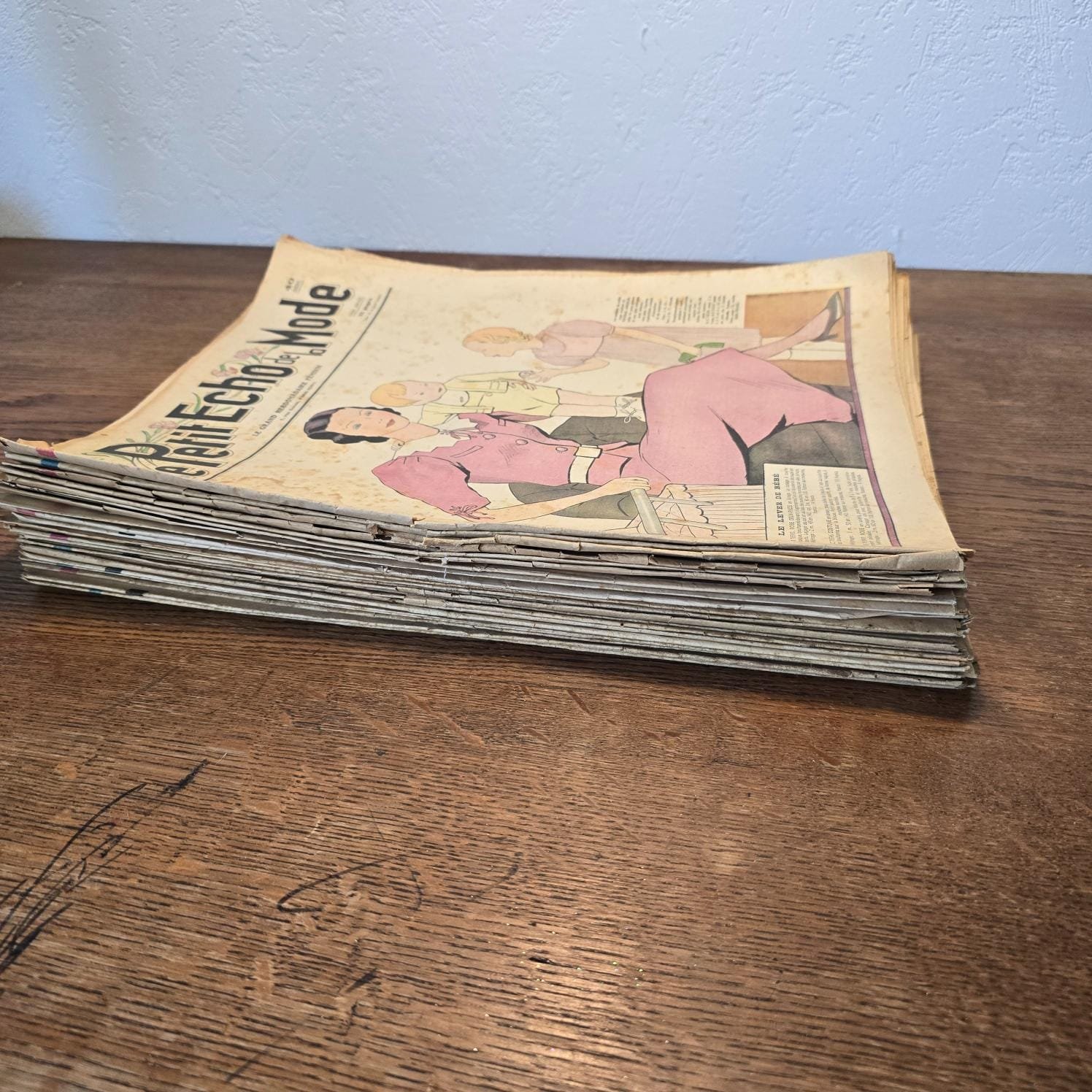 Lot de 30 magazines « Le Petit Écho de la Mode » de 1936 posé en tas sur une table en bois, vue de profil