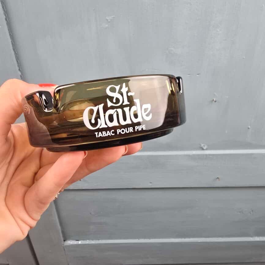 Cendrier publicitaire St Claude en verre fumé tenu à la main, vue du logo blanc