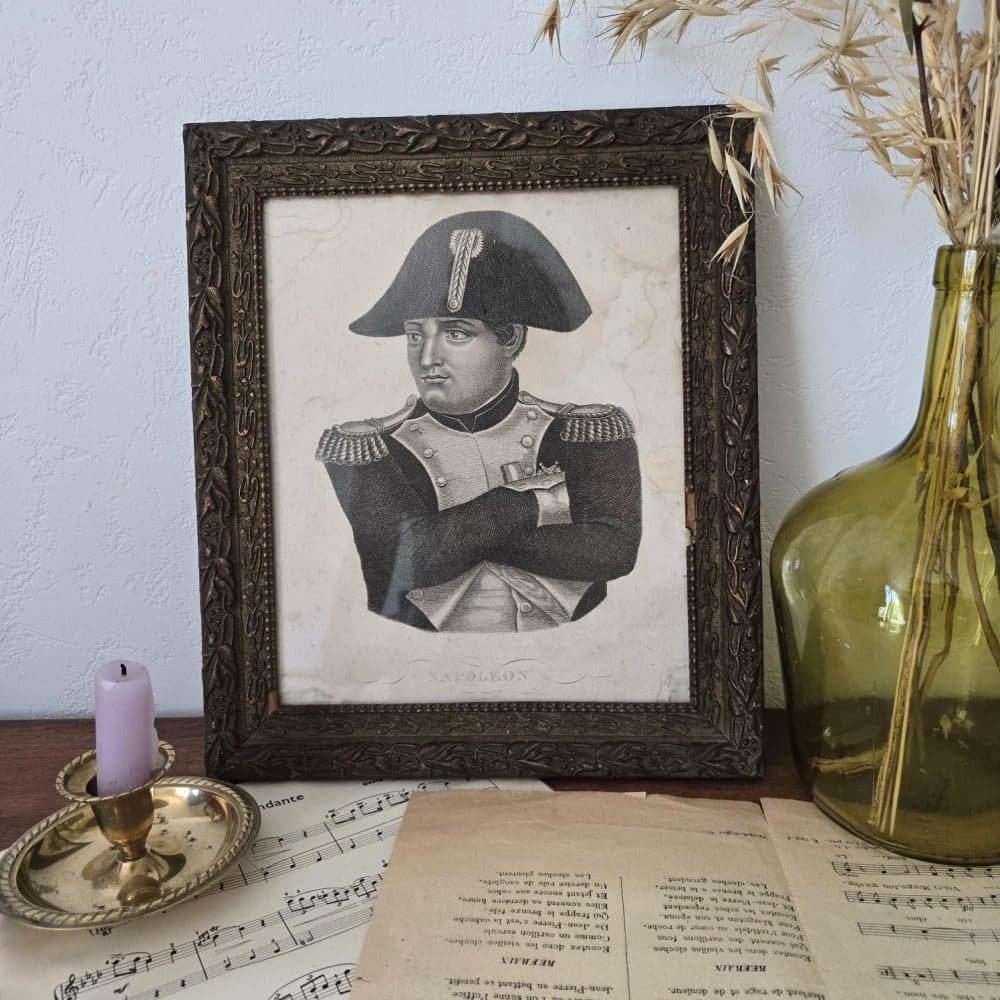 Portrait de Napoléon dans un cadre ancien exposé sur une mise en scène poétique avec partitions anciennes, chandelier et dame-jeanne ambré.