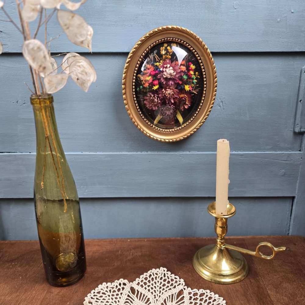 Cadre ancien ovale à verre bombé avec fleurs séchées, accroché au mur dans une mise en scène vintage sur fond bleu.