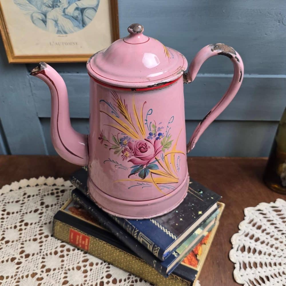 Cafetière ancienne émaillée rose décor fleuri peint à la main, posée sur livres anciens et napperon crochet.