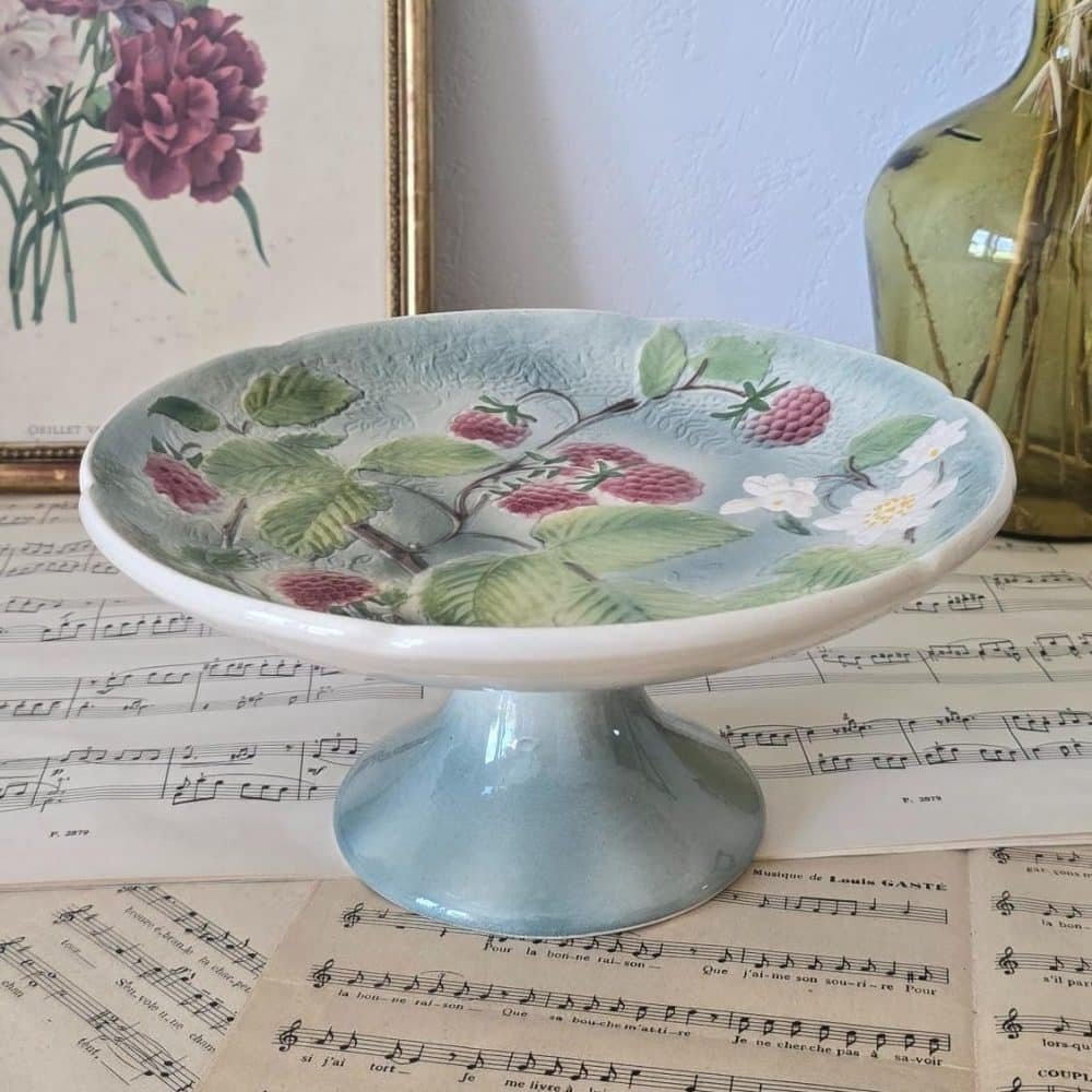 Compotier ancien en barbotine de St Clément, décor framboises et fleurs sur fond bleu, présenté sur partitions anciennes.