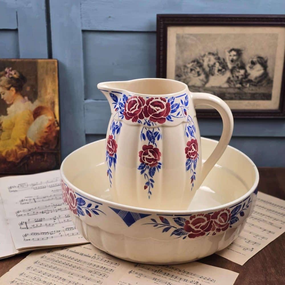 Broc et bassine anciens en faïence K&G Luneville modèle Les Roses, décor floral rouge et bleu, posés sur fond bois et partitions anciennes.