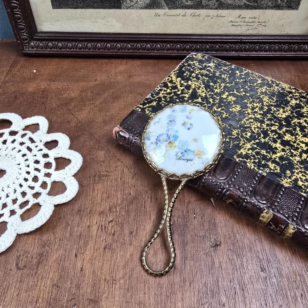 Miroir de sac Limoges posé sur un livre ancien, décor fleuri bleu et jaune.