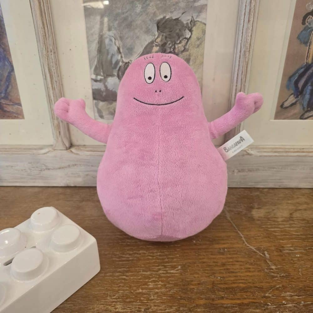 Peluche Barbapapa rose souriante posée sur un meuble en bois, en très bon état, décor mural artistique en fond.