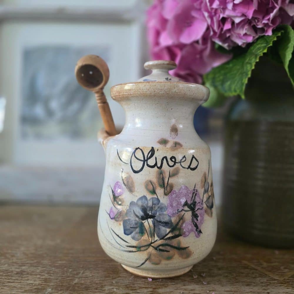 Le pot à olives en grès présenté avec sa cuillère en bois perforée insérée dans le bec latéral.