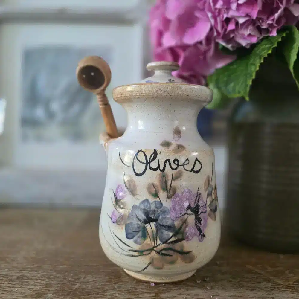 Le pot à olives en grès présenté avec sa cuillère en bois perforée insérée dans le bec latéral.