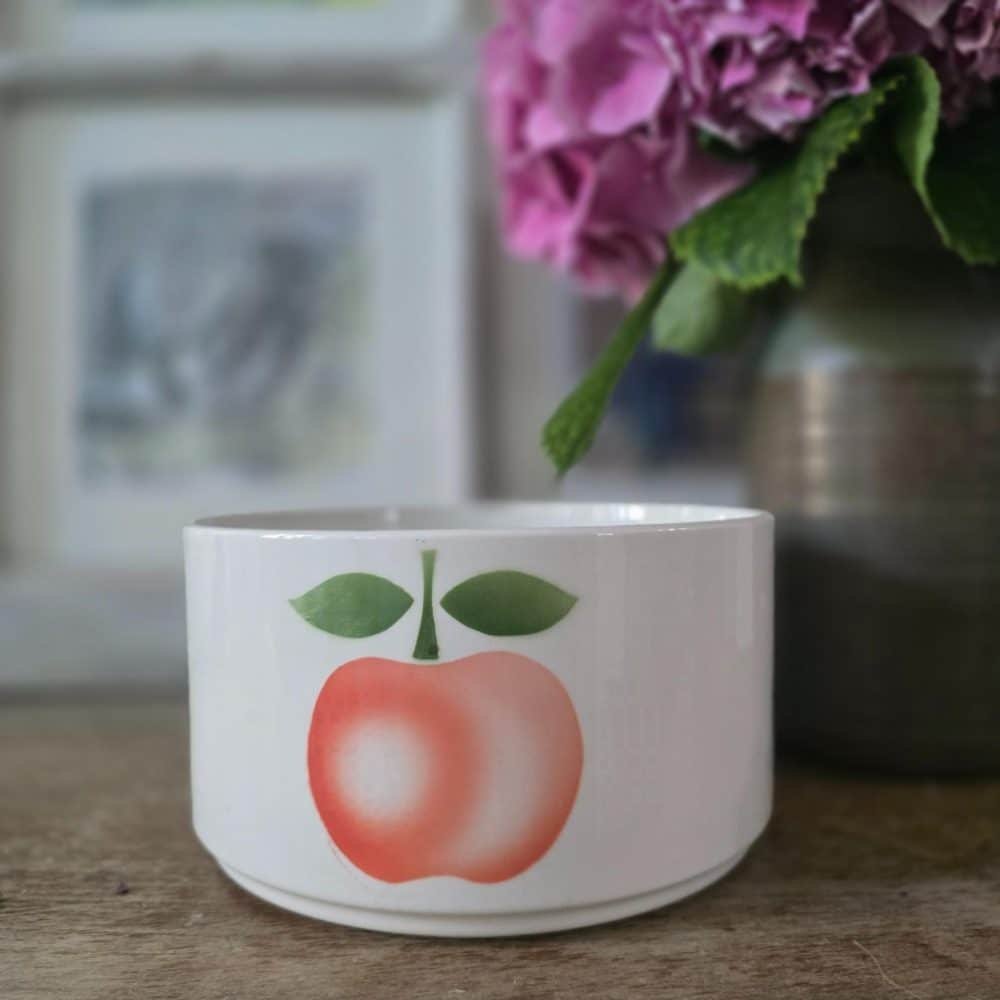 Saladier blanc en faïence Moulin des Loups décoré d’une pomme rose et verte, présenté sur une table en bois devant un vase d’hortensias.